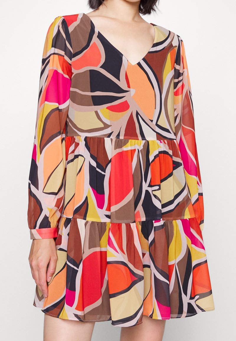 Femme portant une robe à manches longues, col en V, à volants avec un motif abstrait vif dans des tons rouges, orange, rose, marron et beige.