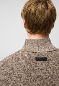 Brązowy sweter z dzianiny z wysokim kołnierzem, z teksturowanym wzorem oraz czarną metką z logo z tyłu przy szyi.