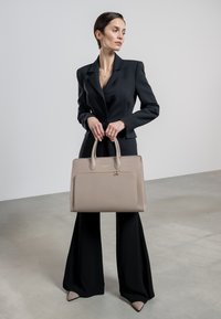 Beige leather handbag with dual top handles, textured surface, and front pocket, held by a person in a fitted black suit and wide-leg pants.