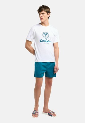 Young man in white graphic T-shirt, teal shorts, and colorful slide sandals standing against a plain light background.