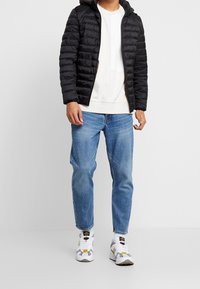 Schwarze Steppjacke mit Kapuze, weißes Sweatshirt, hellblaue Jeans und gemusterte graue Sneaker mit gelben Akzenten. Lässiger, sportlicher Look.
