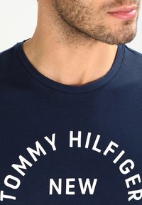 Námořnická modrá bavlněná tričko s kulatým výstřihem, s výrazným bílým textem: "TOMMY HILFIGER NEW" v kruhovém designu na přední straně.