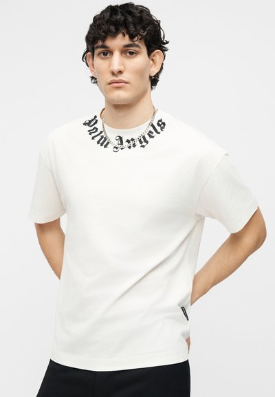 Νέος άντρας με σγουρά μαλλιά φοράει λευκό t-shirt με μαύρο κείμενο "Palm Angels" γύρω από το λαιμό και ασημένιο κολιέ αλυσίδα.