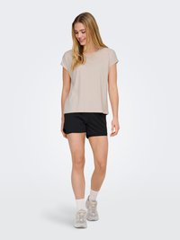 Jeune femme aux longs cheveux blonds vêtue d'un haut beige à manches courtes, d'un short noir, de chaussettes blanches et de baskets grises, debout à l'intérieur.