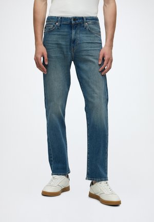 Mand iført blå rette jeans, hvide lædersneakers med brune såler og en hvid skjorte, der står mod en ensfarvet baggrund.