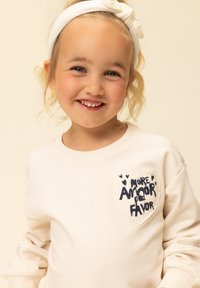 Roomkleurige sweatshirt met een ronde halslijn en lange mouwen, voorzien van een hartvormige patch met marineblauwe tekst "More Amor por Favor." Soepele textuur.