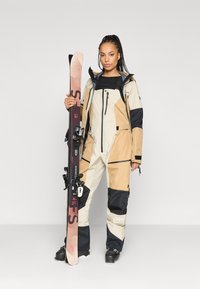 Beige, barna és fekete síruha, struktúrált felülettel. Állítható mandzsettákkal, több zsebbel és kapucnival rendelkezik. Egy sílécet tart a kezében.