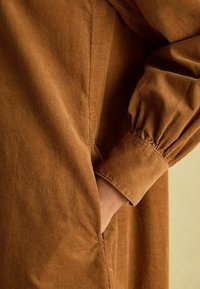 Braunes Cordkleid mit Puffärmeln, ausgestattet mit einer Seiten Tasche und sichtbaren Nähten, welches den strukturierten Stoff und den lockeren Schnitt betont.