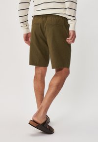 Olivové zelené šortky s volným střihem, elastickým pasem a dvěma zadními kapsami; nošeny s tmavými sandály. Textura se zdá být hladká.