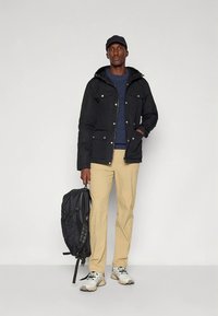 Juoda striukė su gobtuvu, dvi priekinės kišenės, dėvima ant mėlyno megztinio. Smėlio spalvos kelnės ir balti sportbačiai, laiko juodą kuprinę.