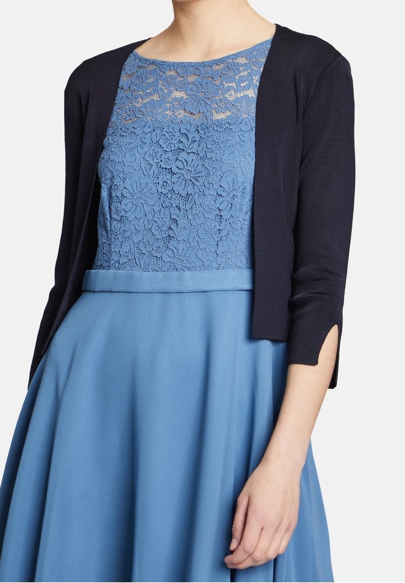 Robe en dentelle bleue avec un cardigan bleu marine. La robe présente un corsage en dentelle florale, des manches courtes et une jupe fluide. Le cardigan a des manches longues et un devant ouvert.