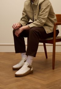 Witte hoge sneakers met bruine accenten, geperforeerd leer en rubberen zolen, gecombineerd met donkerbruine corduroy broek en witte enkelsokken.