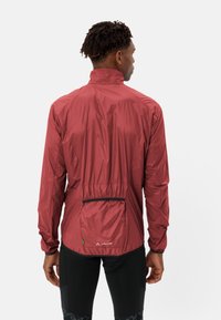 Uomo che indossa una giacca da ciclismo leggera rossa e pantaloni corti da ciclismo neri imbottiti, rivolto di spalle contro uno sfondo chiaro e semplice.