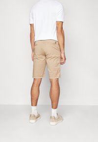 Mann trägt beige knielange Shorts mit Gesäßtaschen und einem kleinen gelben Logo, weiße Socken, beige Sneakers und ein weißes Kurzarmhemd.