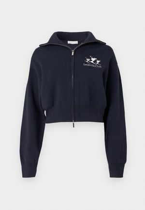 Tumesinine lühendatud lukuga dressipluus kõrge kraega, millel on soonilised varruka- ja vöökohad ning vasakul rinnal valge tikandiga logo.