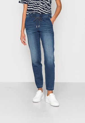 Personne portant un pantalon jogger en denim bleu avec des poignets élastiques et une taille à cordon, associé à des baskets blanches et une chemise à motif noir et blanc.
