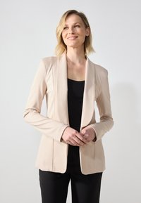 Beige szabott blézer, sima felülettel, nyitott elülső résszel és hosszú ujjakkal, fekete toppal párosítva. Minimalista dizájn, strukturált illeszkedés.