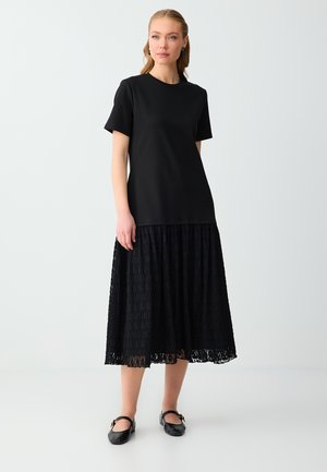 Kobieta o blond włosach w czarnej bluzce z krótkim rękawem, czarnej koronkowej spódnicy midi i czarnych butach Mary Jane stoi na tle jednolitego jasnego tła.