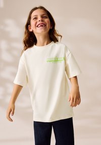 Cremefarbenes T-Shirt mit kurzen Ärmeln, vorne mit einem grünen "Minecraft Movie"-Motiv. Kombiniert mit einer dunklen Jeanshose.