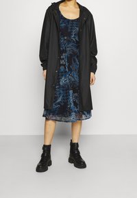 Manteau noir imperméable sur une robe bleue à motif avec des designs floraux. Comprend une capuche, un fermeture à pression et des bottes de combat noires.