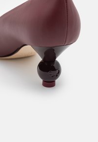 Chaussure à talon haut en cuir bordeaux avec un talon accent noir arrondi arborant un logo, mettant en avant une texture lisse et élégante.