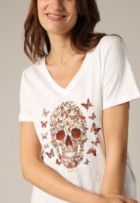 T-shirt en coton blanc à col en V présentant un design de crâne composé de motifs de papillons colorés avec des accents rouges et un texturage détaillé.