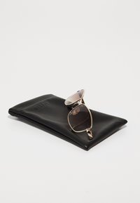 Funda de gafas de sol de cuero negro con logo grabado que sostiene gafas de sol con montura dorada que presentan lentes con degradado rosa y lentes oscurecidos.