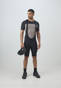 Cuissard de vélo noir et gris avec un panneau en mesh, des manches courtes et des bretelles. Le modèle tient un casque noir et porte des chaussures et des chaussettes foncées.