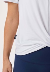 Personne portant une chemise blanche à manches courtes et un pantalon bleu marine, montrant l'étiquette latérale sur la chemise près de la hanche.