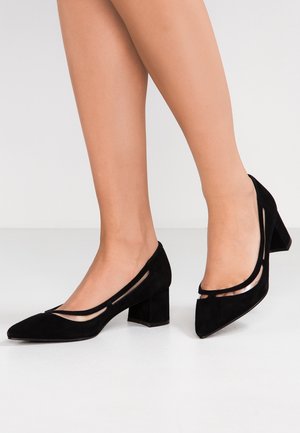 Vrouw die zwarte suède blokhakschoenen draagt met puntige neuzen en transparante meshdetails op een witte vloer.