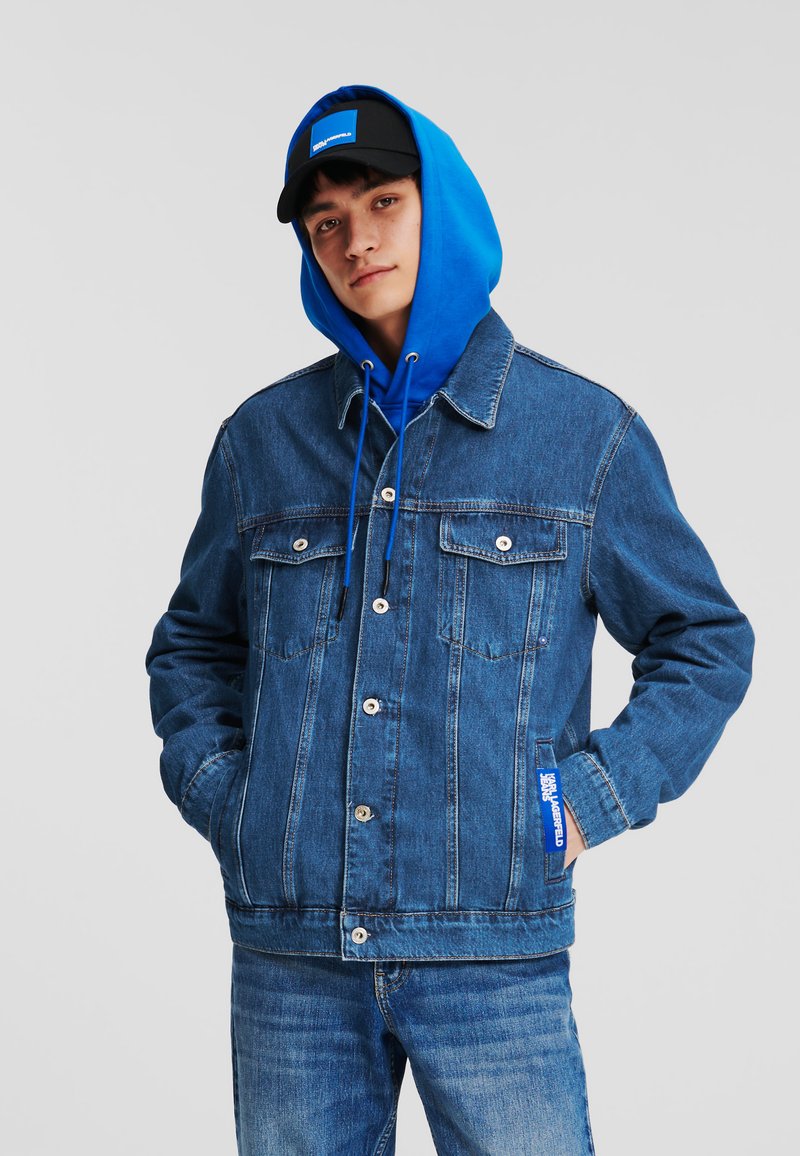 Jachetă din denim de culoare medie, cu două buzunare pe piept, nasturi argintii și un hanorac albastru dedesubt. Asortată cu blugi albaștri și o șapcă neagră.