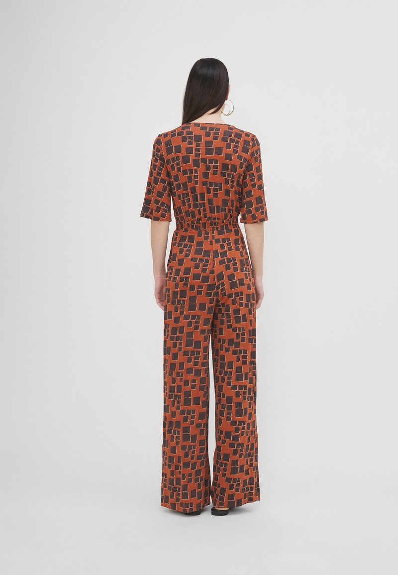 Femme aux longs cheveux noirs portant une combinaison ample à motifs marron et noir, debout de dos sur un fond blanc uni.