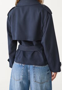 Marineblauwe trenchcoat met een cropped achterkant, voorzien van een ceintuur en een kraag met knopen. Gecombineerd met lichtblauwe, hooggetailleerde jeans.