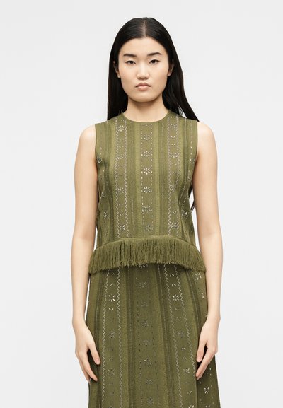 Νέα γυναίκα με μακριά μαύρα μαλλιά που φορά αμάνικη ελιά πράσινη πλεκτή μπλούζα με κρόσσια και διακοσμητικές κάθετες ρίγες, στέκεται μπροστά σε απλό φόντο.