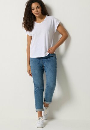 Femme aux cheveux bouclés portant une chemise blanche à manches courtes, un jean bleu retroussé et des baskets blanches, debout contre un mur uni.