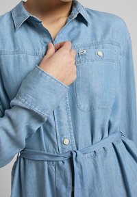 Henkilö, joka pitää päällään vaaleansinistä farkkumekkoa, jossa on napitettu rintatasku ja sidottu vyö, pitäen kaulusta lähellä kaulaa.