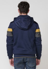 Sudadera con capucha en color navy con acentos en gris y amarillo, mangas texturizadas, puños y dobladillo acanalados. Cuenta con una capucha forrada y un ajuste casual.