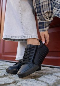 Czarne sneakersy z teksturowanym, sztucznym skórzanym cholewką, płaskimi sznurówkami i gumową podeszwą. Połączone z jasnoszarymi spodniami w szerokim kroju i koszulą w kratę.