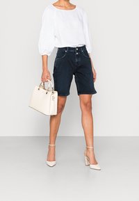 Blouse blanche ample avec des manches bouffantes, short en denim foncé, sac à main beige et chaussures à talons beiges avec des accents texturés.