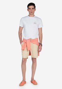 Camiseta blanca con estampado gráfico, sudadera melocotón atada a la cintura, pantalones cortos beige y espadrillas melocotón.