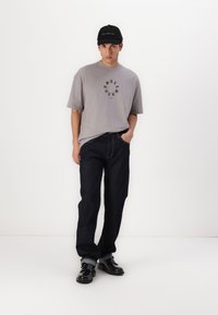 Camiseta gris de gran tamaño con gráfico circular negro, jeans oscuros de pierna recta y zapatos negros combinados con una gorra negra.