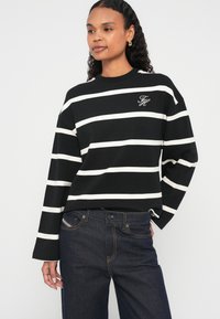 Chemise à manches longues rayée noir et blanc avec un col rond et un logo brodé sur le devant, associée à un jean en denim bleu foncé.