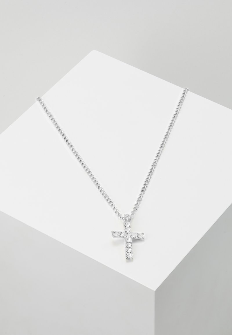 Collier chaîne en argent avec un pendentif croix incrusté de strass clairs, présenté sur une surface géométrique blanche.