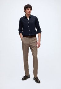 Homme portant une chemise noire à boutons avec les manches retroussées, un pantalon beige, une ceinture marron et des mocassins marron foncé, debout devant un fond uni.