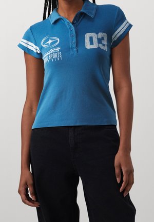 Vrouw draagt een blauw poloshirt met korte mouwen, witte strepen, de tekst "03" en de opdruk "Sports", gecombineerd met een zwarte broek.