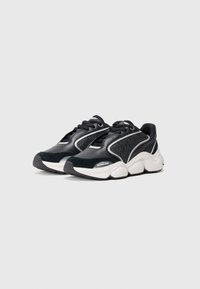 Une paire de baskets noires et blanches avec des semelles épaisses, un design à lacets, et des panneaux latéraux texturés présentant un motif subtil et le nom de la marque.