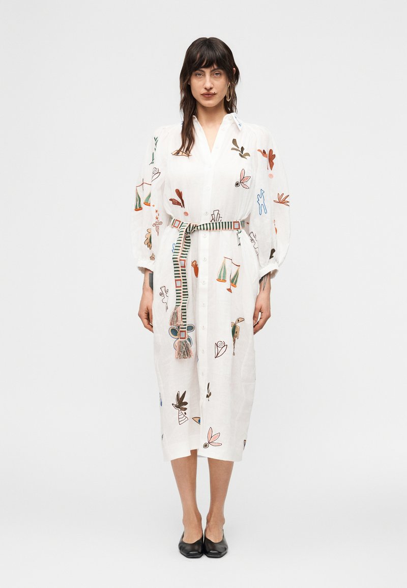 Γυναίκα που φοράει λευκό midi φόρεμα με πολύχρωμη αφηρημένη κεντητική, φουσκωτά μανίκια, ριγέ ζώνη και μαύρα πάνινα παπούτσια, στέκεται μπροστά σε λευκό φόντο.