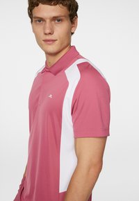 Pikétröja i rosa med vita sidopaneler, korta ärmar och krage. Tillverkad av ett mjukt tyg och prydd med en diskret logotyp på bröstet.