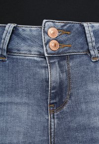Jean en denim bleu clair avec une taille haute, deux boutons en cuivre et des coutures visibles avec une texture délavée.