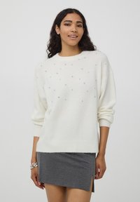 Biały dziany sweter o luźnym kroju, zdobiony rozmieszczonymi akcentami z kryształków i ściągaczami na mankietach. Modelka łączy go z szarą spódnicą.